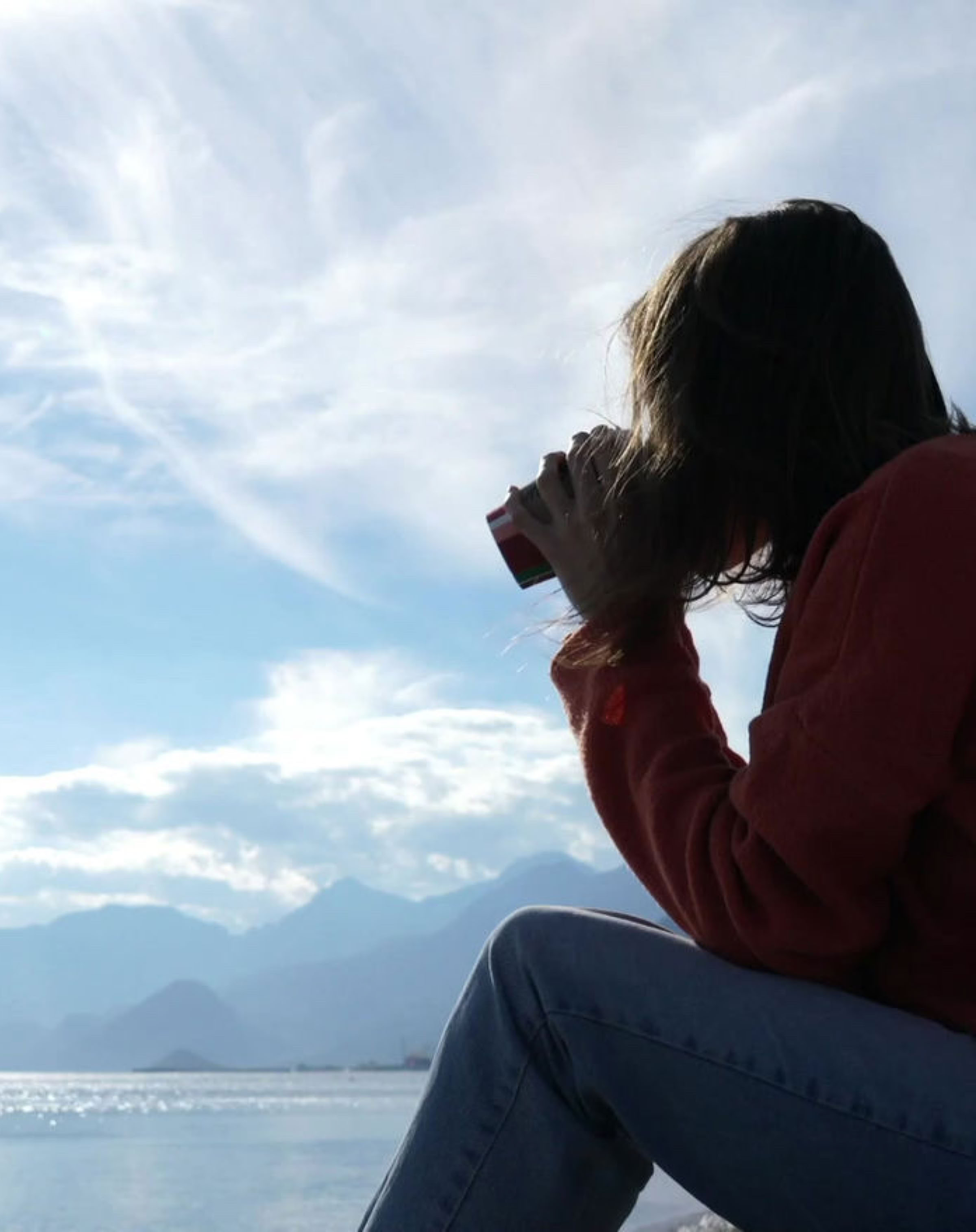 Thoughtful person sitting by the water, holding a drink, with mountains and a cloudy sky in the background.